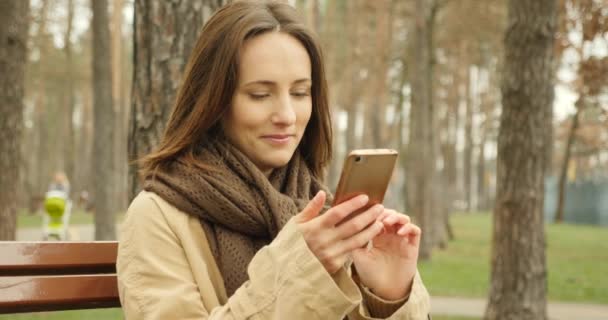 Attractive girl with cellphone scrolling and typing on touch screen while socializing in social networks online wearing brown scarf in autumn park. - Video