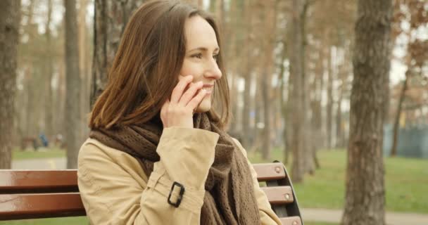Portrét ženy mluvící po telefonu sedí na lavičce v podzimním parku v hnědé šále těší konverzaci. - Záběry, video