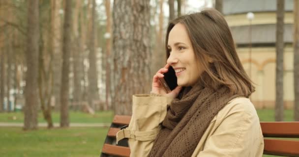 jong brunette vrouw praten op telefoon terwijl zitten op een bank in de herfst park dragen bruin sjaal. - Video