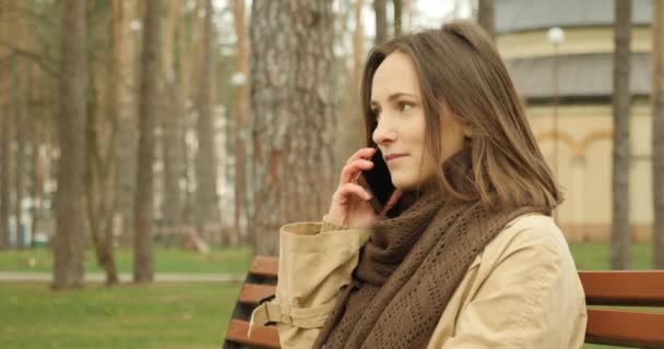Attractive girl starts talking on the phone and laughing and smiling while having a conversation in autumn park on the bench. - Filmati, video
