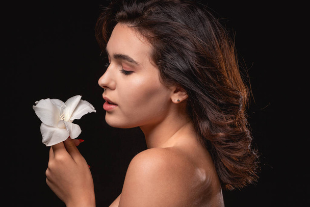 Όμορφη γυναίκα με φωτεινό μακιγιάζ ποζάρουν απομονώνονται σε μαύρο φόντο με λευκό λουλούδι. - Φωτογραφία, εικόνα