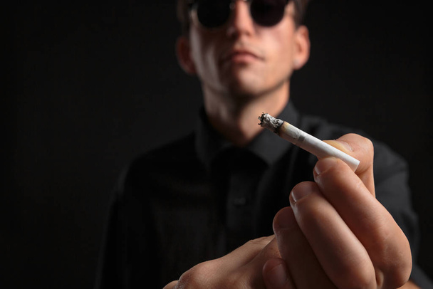 Nahaufnahme eines stylischen jungen Mannes in schwarzem T-Shirt und Sonnenbrille mit Zigarette - Foto, Bild