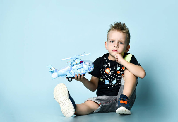 Pozytywne blond chłopiec w stylowej odzieży casual siedzi na podłodze i trzymając helikopter zabawka obecny w ręku nad niebieskim tle - Zdjęcie, obraz