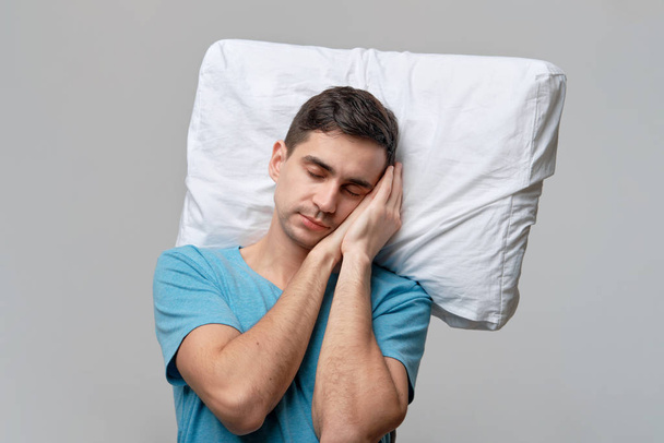 Tired brunet man in a blue tee resting on a white pillow isolated over grey background. - Foto, Imagem
