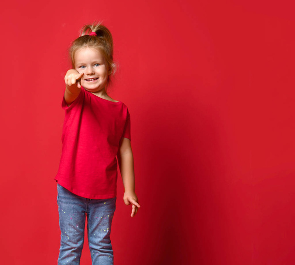 Small happy excited blond girl in stylish casual clothing standing and pointing at camera over red wall background - 写真・画像