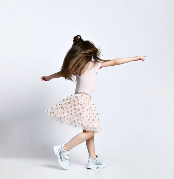 Small girl in casual summer dress and white sneakers standing backwards and moving over white wall background - Fotó, kép