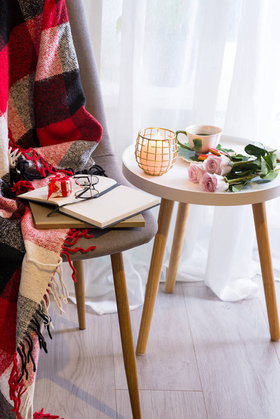 Masanın üzerinde pembe güller olan bir mum pencerenin yanında bir sandalye, bir ekose ve bir kitapla birlikte. - Fotoğraf, Görsel