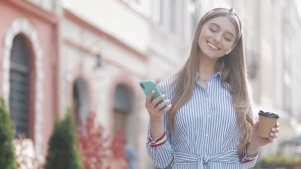 Young Smiling Woman with Brown Hair Wearing Stylish Striped Shirt and Headband Looking Pleased and Happy Using her Smartphone and Holding Coffee Cup. Girl Walking at the Old City Street. - Video