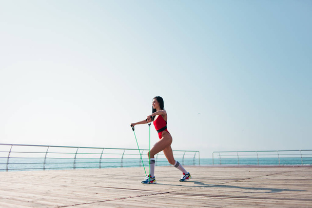 Młoda kobieta w czerwonym sporcie body robi ćwiczenia aerobowe z ekspanderem na plaży rano - Zdjęcie, obraz