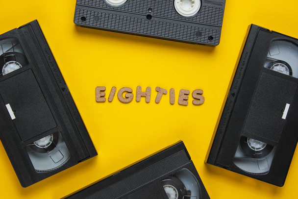Conceito de estilo retrô, anos 80. Vídeo cassete em um fundo amarelo com a palavra Oitenta de letras de madeira. Vista superior, minimalismo - Foto, Imagem