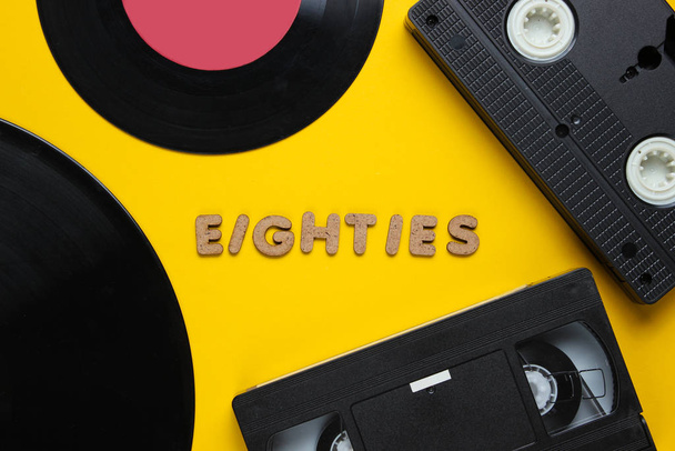 Retro style concept, 80s. Video cassette and vinyl records on yellow background with the word Eighties from wooden letters. Top view, minimalism - Photo, Image