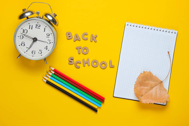 Okul zamanı minimum konsept. Not defteri, renkli kalemler, sarı arkaplandaki çalar saat ve okula tahta harflerle dönüş sloganı. Üst görünüm - Fotoğraf, Görsel
