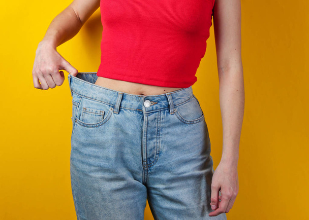 減量の概念。黄色の背景に大きな青いジーンズをしようとしている女性 - 写真・画像