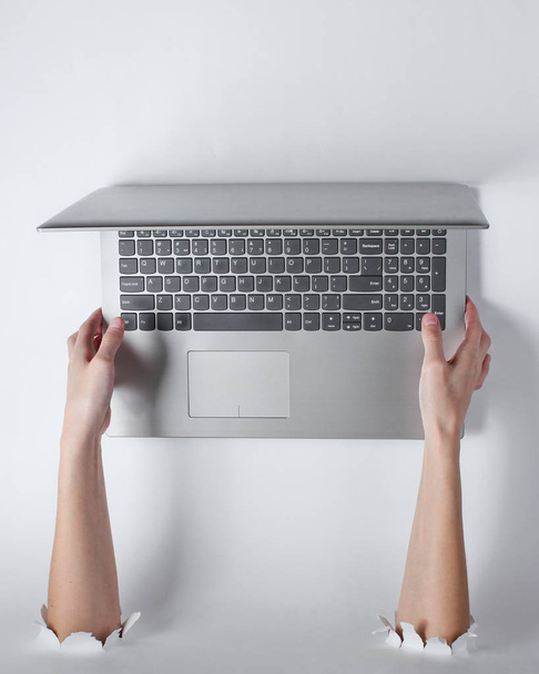 Mãos femininas segurando um laptop através dos orifícios rasgados do fundo branco. Arte empresarial criativa
 - Foto, Imagem