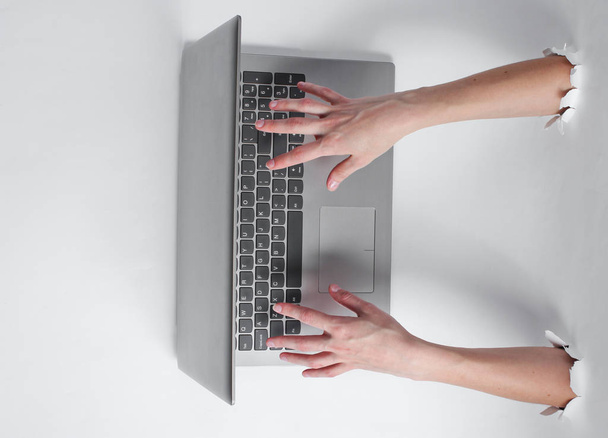 Female hands are typing on laptop keyboard through the torn holes of white background. Creative business art - 写真・画像