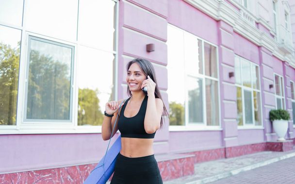 Vrolijke sportieve vrouw in sportschoeisel gaat voor training met yoga mat en praten aan de telefoon buiten tegen de achtergrond van het gebouw - Foto, afbeelding