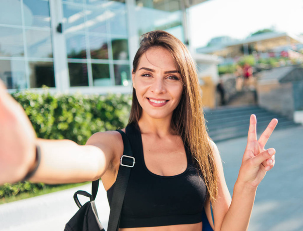 Selfie portret Młoda, wesoła kobieta w sportowej bluzce z torbą na ramionach uśmiechnięta i pokazująca symbol V na zewnątrz - Zdjęcie, obraz
