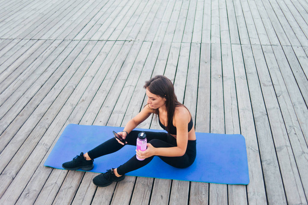 Mujer sana alegre de la aptitud en ropa deportiva sentada en la estera del yoga y charlando en el teléfono inteligente al aire libre en tableros de madera
 - Foto, Imagen