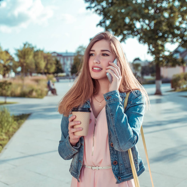 beautiful girl in denim jacket, in summer in city, calls by phone, listens to voice message, an online application on Internet. In hand is cup of hot coffee tea. Background road trees. - Фото, зображення
