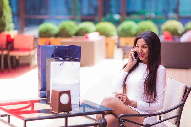 Young smiling afro woman talking on the phone while sitting in an outdoor cafe with shopping bags on the table - Foto, imagen