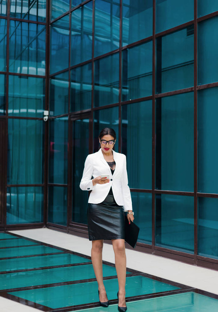 Mujer afro de negocios profesional en elegante chaqueta blanca y falda con carpeta con documentos en sus manos use teléfono inteligente contra el fondo del centro de negocios con ventanas azules
 - Foto, imagen