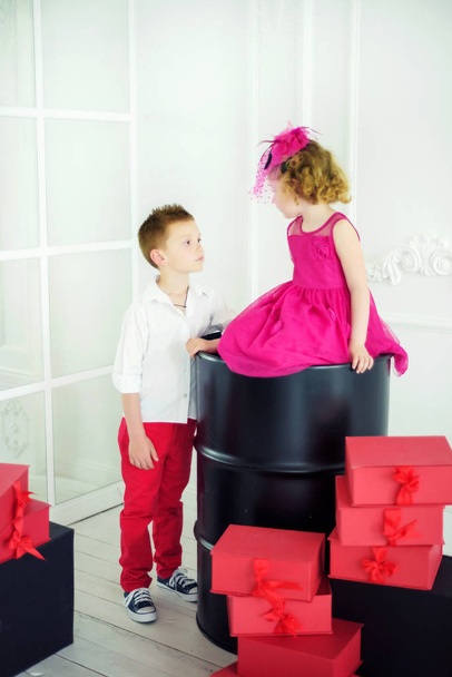 Маленька 4-річна дівчинка в красивій рожевій сукні і рожевий капелюх з вуаллю сидить на чорному бочці, дивлячись на хлопчика в білій сорочці і червоні кинджали з подарунковими коробками навколо. Брат і сестра на день народження
 - Фото, зображення