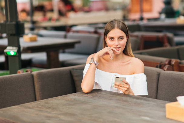 Modieuze jonge vrouw die een smartphone in haar handen houdt terwijl ze aan een tafel zit in een café - Foto, afbeelding