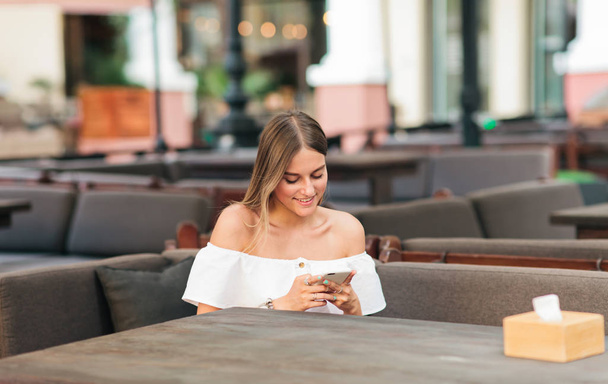 Mujer rubia feliz sosteniendo teléfono inteligente en sus manos mientras se sienta en una mesa en la cafetería de la calle
 - Foto, Imagen