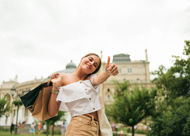 Alegre hermosa mujer adicta a las compras con bolsas de papel y muestra el pulgar hacia arriba y posando en la cámara al aire libre
 - Foto, imagen