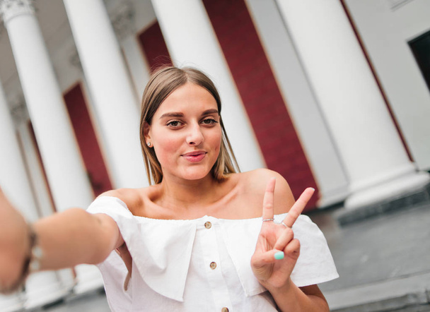 Selfie πορτρέτο μιας χαρούμενης χαρισματικής γυναίκας δείχνει χειρονομία v στο φόντο ενός κτιρίου με κίονες στην εξωτερική - Φωτογραφία, εικόνα