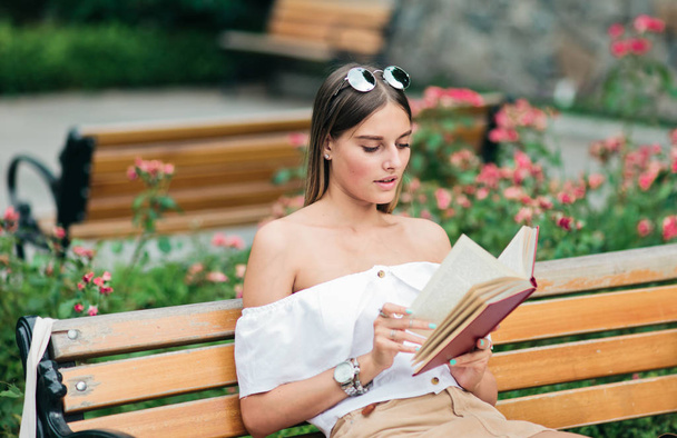 Fiatal szőke nő olvas egy könyvet lelkesedéssel, miközben ül egy padon egy parkban - Fotó, kép