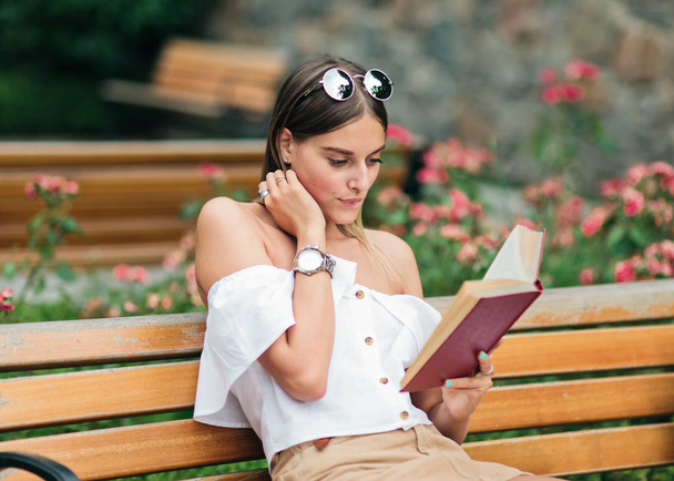 Блондинка-красавица с энтузиазмом читает книгу, сидя на скамейке в парке
 - Фото, изображение