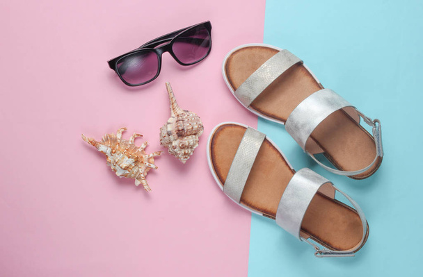 Креативный летний пляж лежал. Кожаные женские сандалии, ракушки, солнечные очки на пастельно-сине-розовом фоне. Вид сверху
 - Фото, изображение