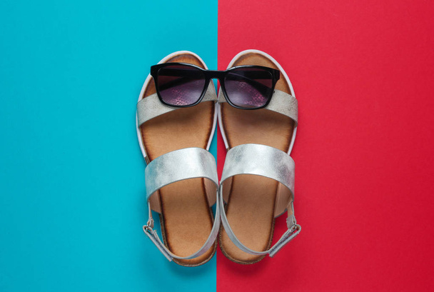Kreatywne lato plaża płaska leżała. Skórzane damskie sandały, okulary przeciwsłoneczne na niebieskim czerwonym tle. Widok z góry - Zdjęcie, obraz