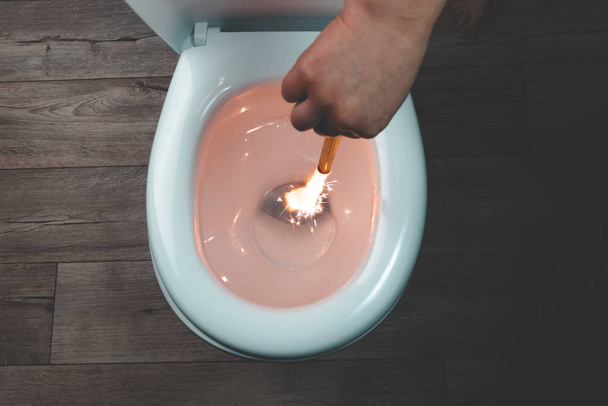 white toilet bowl on a wooden background. the hand lowers the lit firecracker. close-up. - Photo, Image