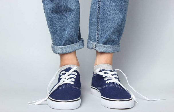 Fashion studio shot. Female legs in jeans and sneakers with untied laces on gray background. - 写真・画像