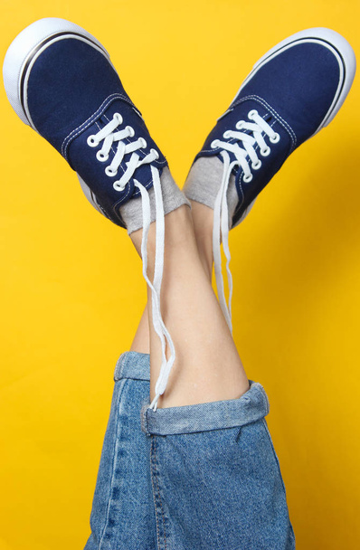 Fashion studio shot. Raised female legs in jeans and sneakers with untied laces on yellow background. - Foto, imagen