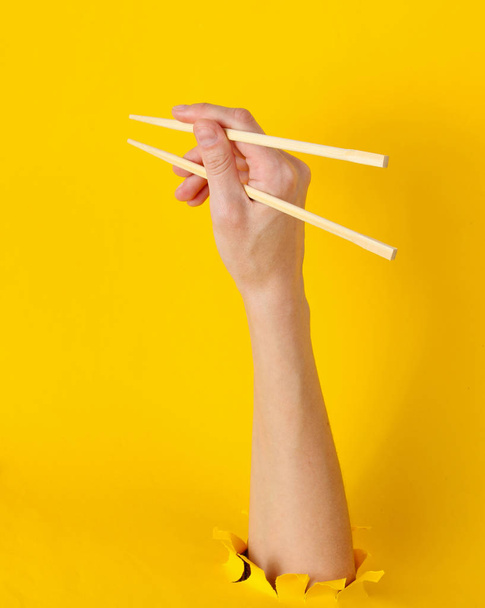黄色い背景に破れた穴から女性の手が箸を持ちます。最小限の食品コンセプト。トップ表示 - 写真・画像