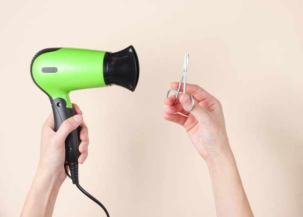Personal care, beauty concept. Female hands holding nail scissors and hairdryer on a beige background. Top view - Photo, Image