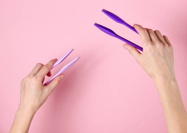 Female hands holding kitchen tweezers on pink background. Top view - Foto, Imagen