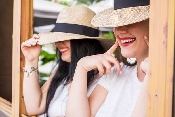 Nahaufnahme Porträt eines jungen kaukasischen Mädchenpaares lächelt und lacht verborgen von modischen Hüten - Freunde haben Spaß zusammen Konzept - trendige moderne Frau mit neuem Lippenstift und glücklichem Lebensstil - Foto, Bild