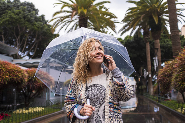 attraktive und fröhliche Mode trendige junge kaukasische Frau mittleren Alters, die mit einem durchsichtigen Regenschirm durch die Stadt läuft - Konzept für die kalte Jahreszeit im Stadtpark - Foto, Bild