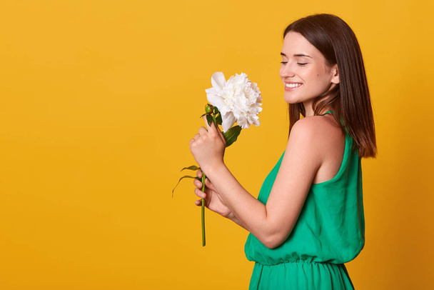 Close up portrait of adorable young woman standing isolated over pink background with charming smile, having happy look, holding white peony flower in hands and smells it. St. Valentines Day. - Фото, изображение