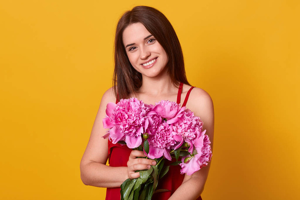 Smiling cute female posing isolated over yellow background in studio, looking directly at camera with charming smile, holding rosy peonies in hands, wearing red dress, having straight dark hair. - Photo, image