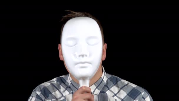 Ein junger Mann nimmt seine Maske ab und zeigt Emotionen im Gesicht. Wütender Mann im Hemd. ein Mann versteckt sein Gesicht hinter einer weißen Maske auf schwarzem Hintergrund - Filmmaterial, Video