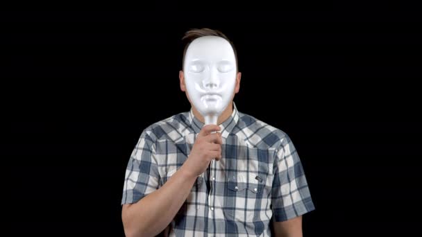 A young man takes off his mask and shows an emotion of surprise on his face. The man stares at the camera in surprise. A man hides his face behind a white mask on a black background. - Footage, Video