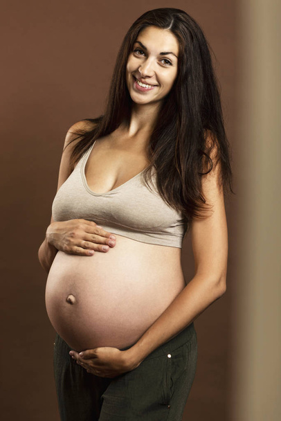 A young pregnant brunette woman smiles and gently touches a large belly. Tenderness and expectation of birth.  - Photo, Image