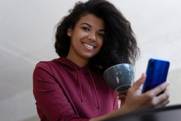 Portret stylowej Afroamerykanki blogerki trzymającej filiżankę kawy i korzystającej ze smartfona, komunikacji, patrzącej w kamerę i uśmiechniętej, siedzącej w nowoczesnej kawiarni. Koncepcja przerwy na kawę - Zdjęcie, obraz