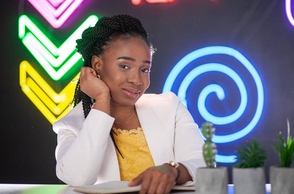 Afro-Amerikaans meisje zittend op een achtergrond van neon tekens op een zwarte achtergrond. Mooie jonge vrouw met kleine staarten. - Foto, afbeelding
