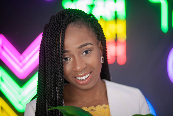 African-American girl sitting on a background of neon signs on a black background. Beautiful young woman with small pigtails. - Foto, Bild